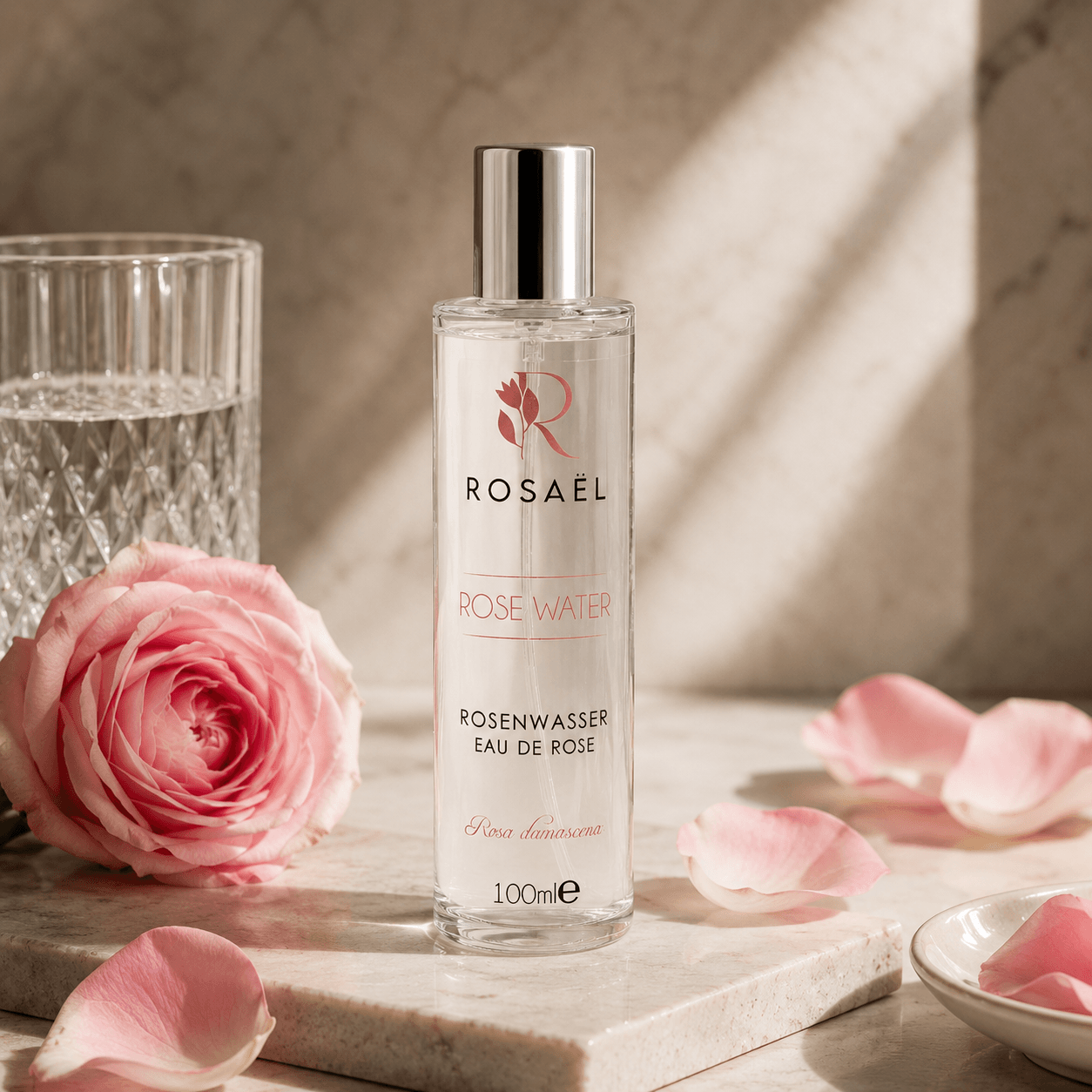 Rosaël Rosenwasser in Glasflasche auf hellem Stein mit Rosenblüte und weichem Morgenlicht