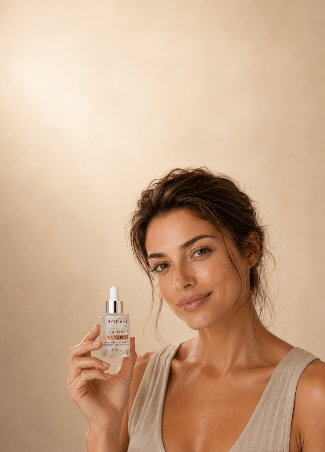 Natürliche Frau hält das kleine Rosaël Serum mit Pipette im Hochformat vor warmem beigem Studiohintergrund.